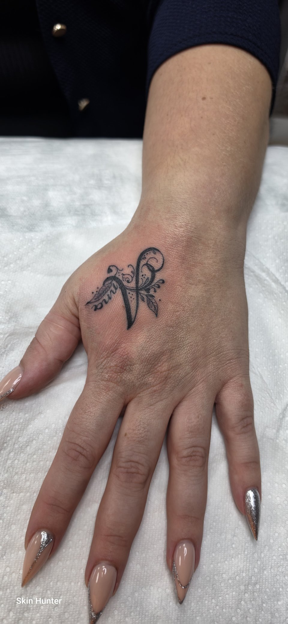 Feiner Buchstabe mit Ranke als kleines Tattoo auf dem Handrücken