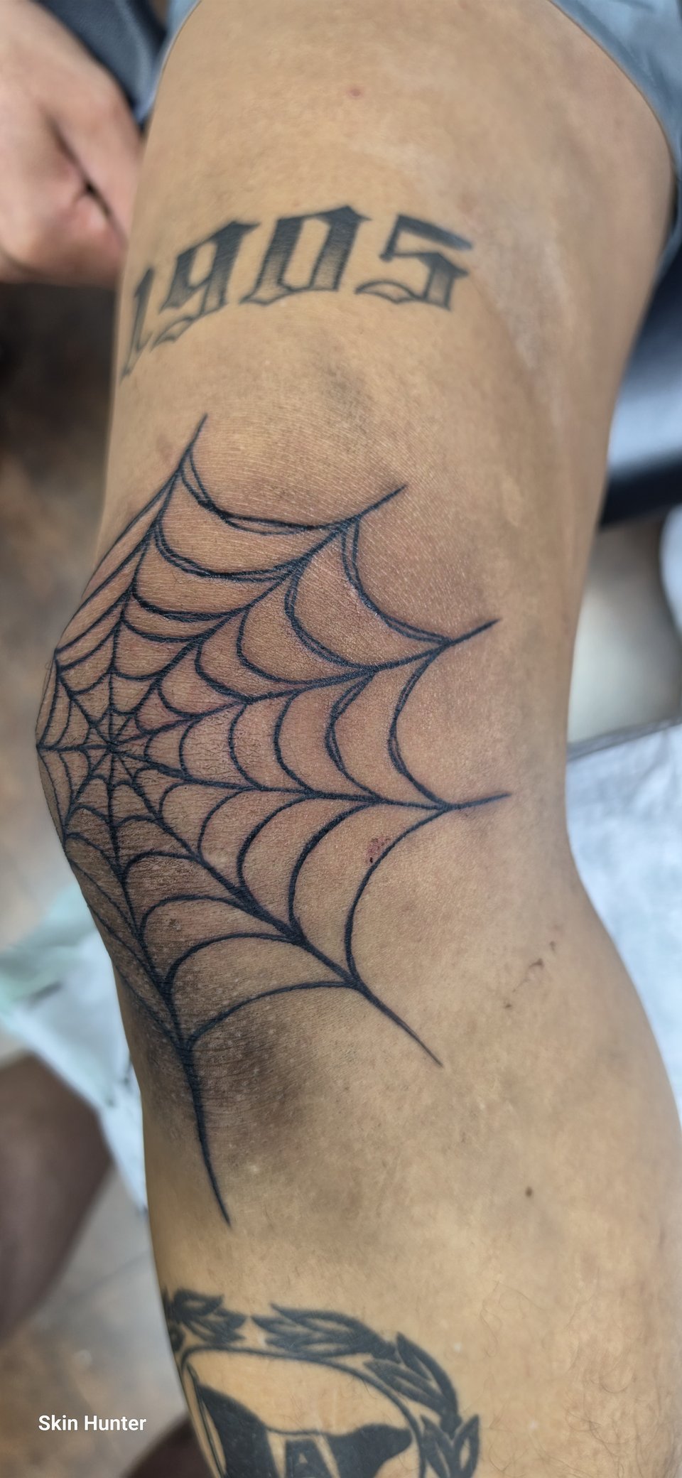 Spinnennetz-Tattoo am Ellenbogen, darüber die Jahreszahl 1905